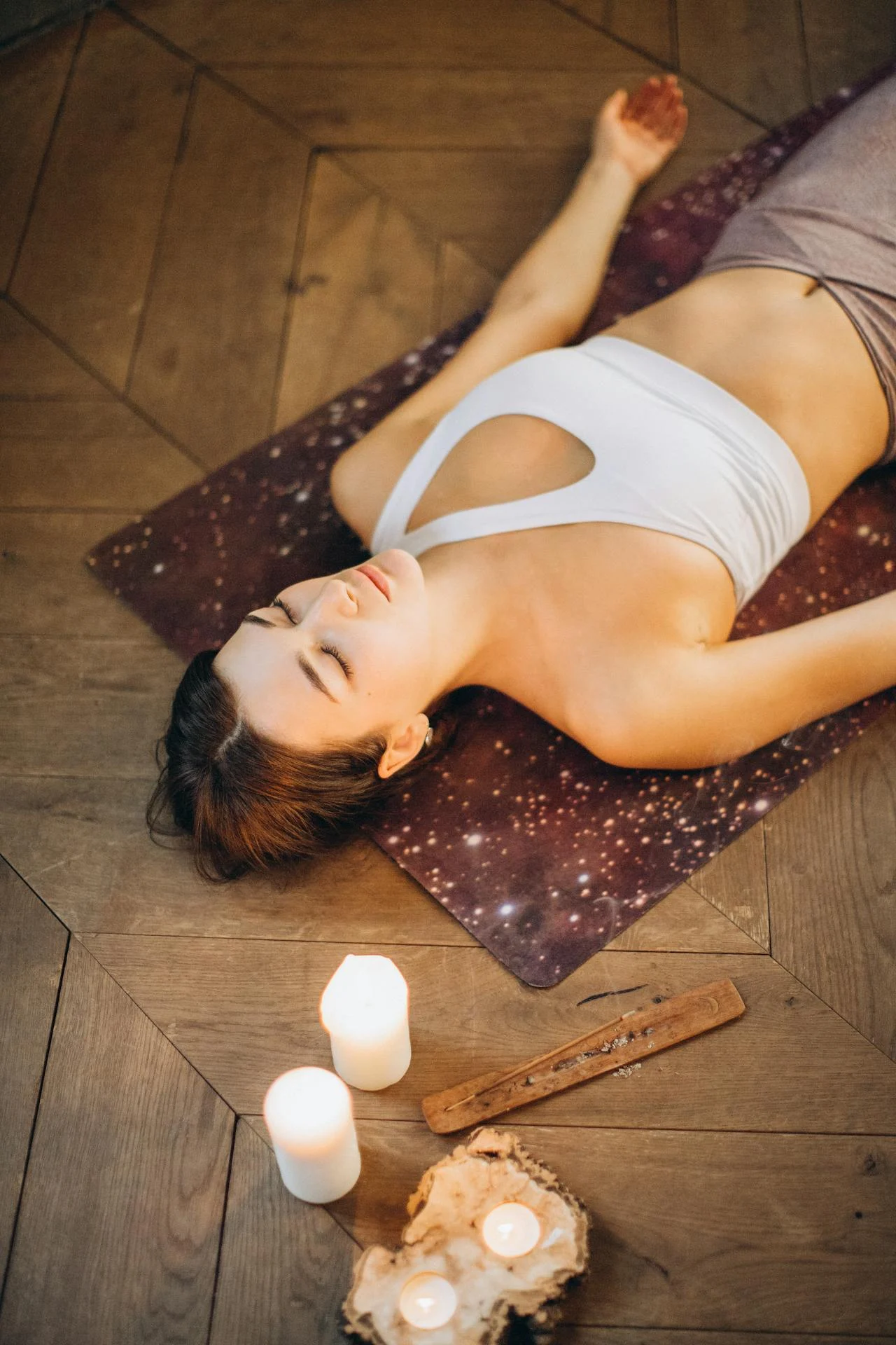 Woman lying in Savasana yoga pose on a mat, surrounded by lit candles and incense, embracing deep relaxation.