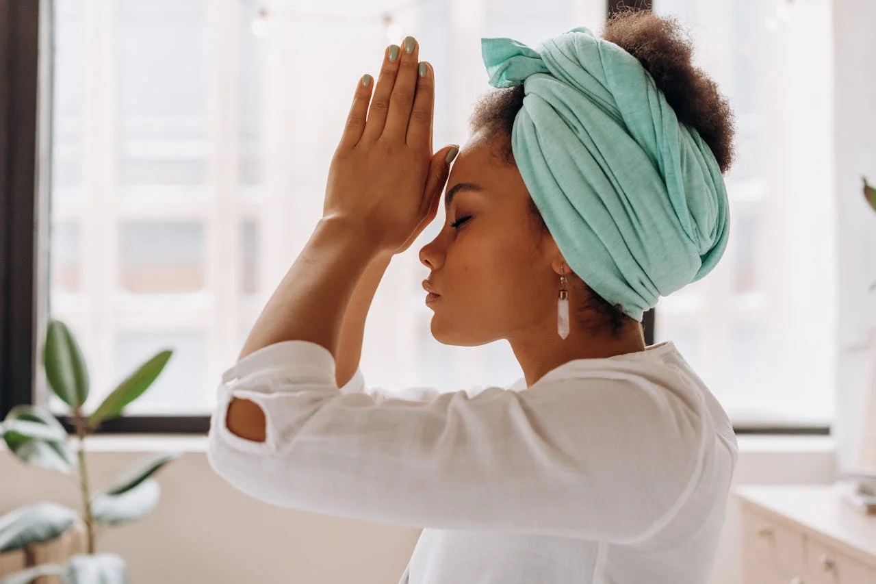 Peaceful woman practicing meditation indoors, with hands in prayer pose and eyes closed, surrounded by natural light.