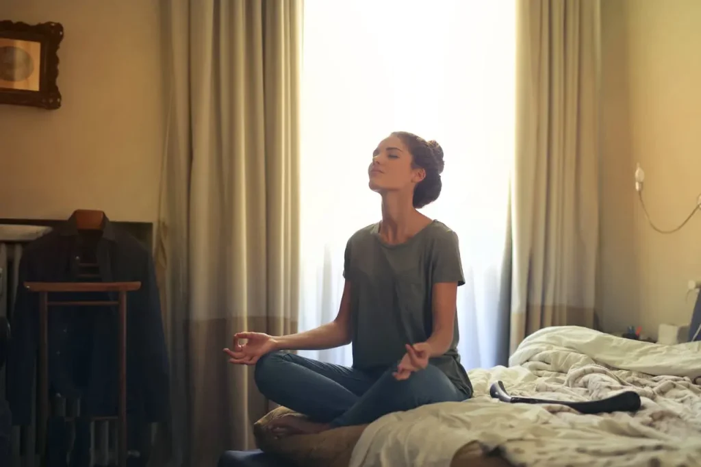Young woman meditating peacefully in a cozy bedroom, sitting cross-legged on the bed with eyes closed and soft morning light streaming through the curtains.