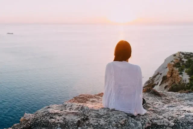 Woman sitting peacefully on a cliff, watching the sunset over the ocean — a quiet moment of reflection and calm.